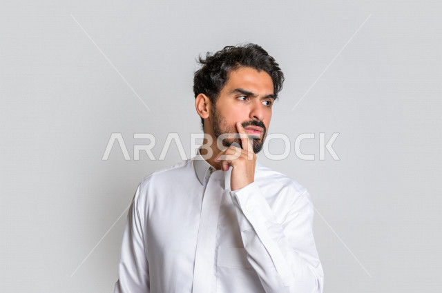 بورتريه لرجل عربي سعودي خليجي باللباس التقليدي , يقوم بإيماءات وجه ويدين تدل على التفكير و الحيرة , خلفية بيضاء .