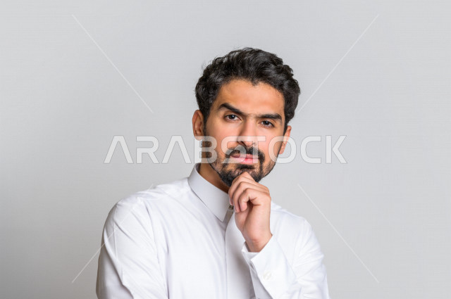 بورتريه لرجل عربي سعودي خليجي باللباس التقليدي , يقوم بإيماءات وجه تدل على التفكير و التركيز ، خلفية بيضاء .