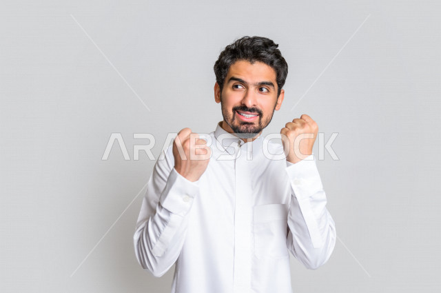 بورتريه لرجل عربي سعودي خليجي باللباس التقليدي , يقوم بإيماءات وجه تدل على السعادة ، إشارات يدين تدل على الفرحة بالفوز ، خلفية بيضاء .
