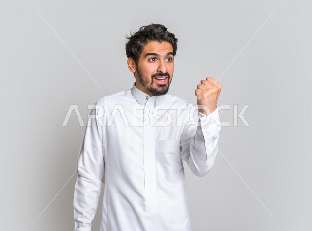 بورتريه لرجل عربي سعودي خليجي باللباس التقليدي , يقوم بإيماءات وجه تدل على السعادة ، إشارات يدين تدل على الفرحة بالفوز ، خلفية بيضاء .