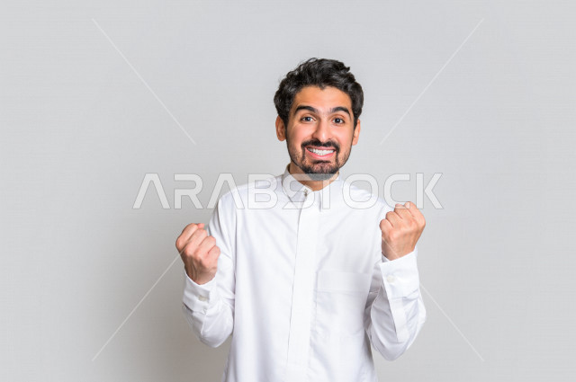 بورتريه لرجل عربي سعودي خليجي باللباس التقليدي , يقوم بإيماءات وجه تدل على السعادة ، إشارات يدين تدل على الفرحة بالفوز ، خلفية بيضاء .