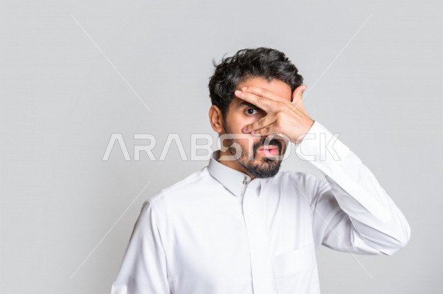 بورتريه لرجل عربي سعودي خليجي باللباس التقليدي , يقوم بإيماءات وجه مختلفة ، إشارة يد تدل على الإختباء و استراق النظر ، خلفية بيضاء .