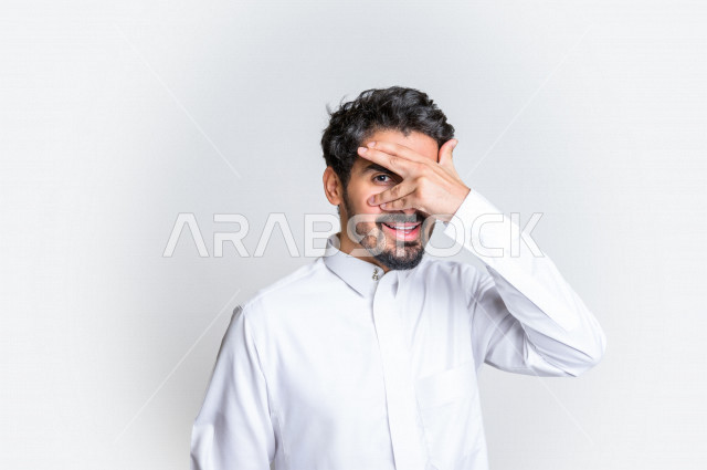 بورتريه لرجل عربي سعودي خليجي باللباس التقليدي , يقوم بإيماءات وجه مختلفة ، إشارة يد تدل على الإختباء و استراق النظر ، خلفية بيضاء .