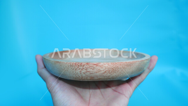 صورة مقربة ليد تمسك بطبق خشبي فارغ، حرف وصناعات يدوية، طبق مصنوع من الخشب، خلفية زرقاء
