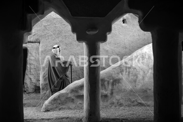 صورة بالابيض والاسود لرجل مسن عربي سعودي خليجي يرتدي الزي السعودي الشعبي التقليدي، زي تراثي للمناسبات الوطنيه والاحتفالية، التجول في إحدى المباني الطينية الشعبية، بيوت تراثية قديمة، جولة بين الأحياء الشعبية