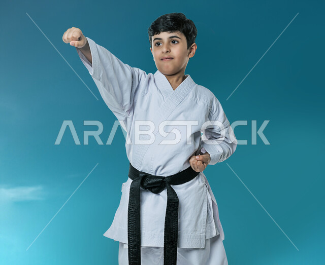 بورتريه لولد عربي خليجي سعودي يمارس رياضة الكاراتيه، زي الكاراتيه الأبيض مع الحزام الأسود، تعلم رياضة التايكواندو، انشطة رياضية ترفيهية للأطفال، رياضة قتالية للدفاع عن النفس، خلفية زرقاء
