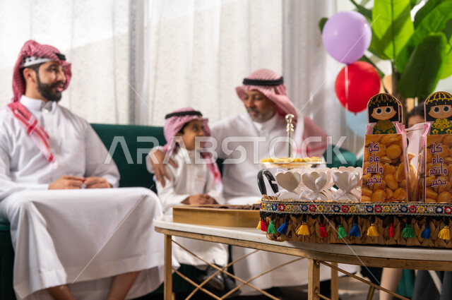 صورة مقربة لتوزيعات العيد وسلة القرقيعان, عائلة عربية خليجية سعودية تحتفل في العيد السعيد, الزيارات والتجمعات و الاحتفالات في المناسبات و الأعياد الاسلامية, بهجة وفرحة العيد, ارتدراء الزي السعودي التقليدي ,ايماءات تدل على الفرح  والسعادة .