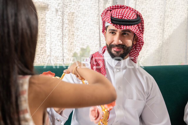 Delight and joy among family and relatives, a close-up of a Gulf Arab young man celebrating the Happy Eid, good reception and honoring the guest, gatherings and visits on Islamic holidays and occasions, gestures indicating joy, wearing the Saudi dress and
