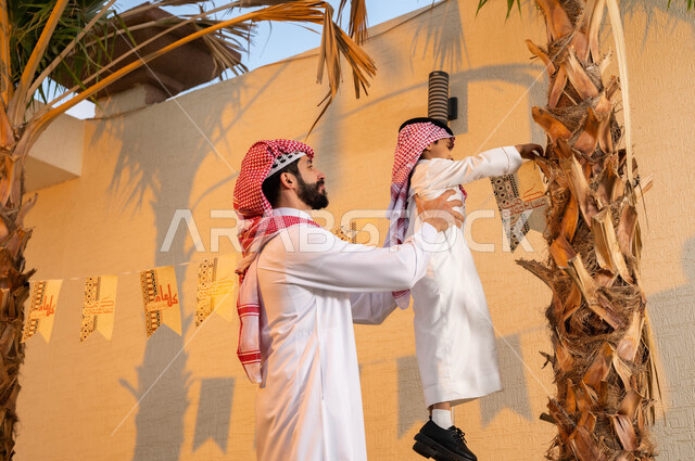 Celebrating the advent of the Eid, wearing the Saudi dress and shemagh, a young Saudi Arabian Gulf man raising his son to put on the decorations for the Happy Eid, decorations and decorations for the Happy Eid, Islamic holidays and occasions.