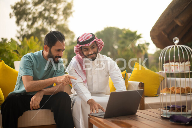 استخدام الحاسوب المحمول والعمل عن بعد،  رجل عربي خليجي سعودي يرتدي الثوب السعودي التقليدي والشماغ السعودي يجلس مع صديقه ويجري اتصال هاتفي، دردشة مع الأصدقاء اجراء مكالمة هاتفية، الجلوس في الهواء الطلق، الطبيعة الخضراء
