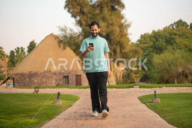 ايماءات وجه تدل على الفرح والسرور ، شاب عربي خليجي سعودي يمسك بيده الهاتف المحمول يقضي أوقات ممتعة في إحدى الحدائق في السعودية، صورة الجسم بالكامل ، نزهه في الهواء الطلق، جولة في الطبيعة الخضراء