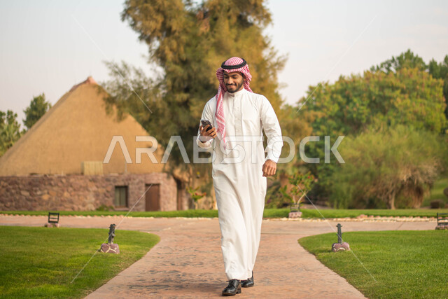 صورة الجسم بالكامل ، شاب عربي خليجي سعودي يرتدي الثوب السعودي التقليدي والشماغ السعودي يمسك بيده الهاتف المحمول يقضي أوقات ممتعة في إحدى الحدائق في السعودية ، نزهه في الهواء الطلق، جولة في الطبيعة الخضراء