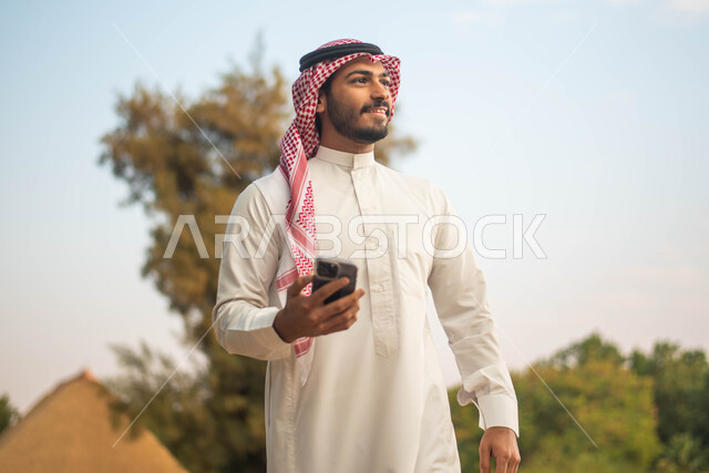 جولة في الطبيعة الخضراء ، رجل عربي خليجي سعودي يرتدي الثوب السعودي التقليدي والشماغ السعودي يمسك بيده الهاتف المحمول يقضي أوقات ممتعة في إحدى الحدائق في السعودية ، نزهه في الهواء الطلق