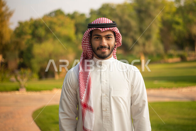 النظر إلى الكاميرا ،  نزهه في الهواء الطلق، جولة في الطبيعة الخضراء ، رجل عربي خليجي سعودي يرتدي الثوب السعودي التقليدي والشماغ السعودي يقضي أوقات ممتعة في إحدى الحدائق في السعودية، التقاط صورة تذكارية