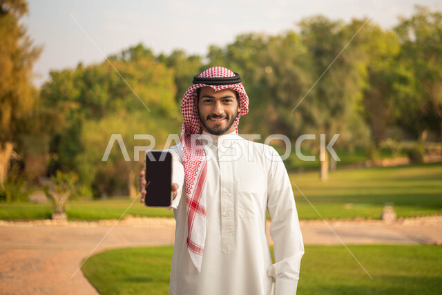 نزهه في الهواء الطلق، عرض شاشة سوداء فارغة ،صورة أمامية لشاب عربي خليجي سعودي يرتدي الزي السعودي التقليدي يمسك بيده الهاتف المحمول يقضي أوقات ممتعة في إحدى الحدائق في السعودية،  جولة في الطبيعة الخضراء