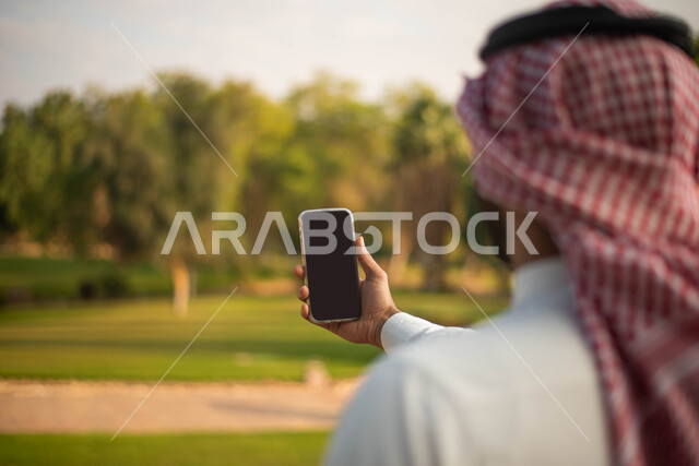  بث مباشر ، نزهه في الهواء الطلق، جولة في الطبيعة الخضراء ، صورة من الخلف لشاب عربي خليجي سعودي يرتدي الزي السعودي التقليدي يقضي أوقات ممتعة في إحدى الحدائق في السعودية، التقاط صور تذكارية من خلال الهاتف المحمول