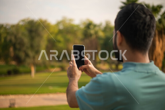 صورة من الخلف لرجل عربي خليجي سعودي  يقضي أوقات ممتعة في إحدى الحدائق في السعودية، التقاط صور تذكارية من خلال الهاتف المحمول، بث مباشر، نزهه في الهواء الطلق، جولة في الطبيعة الخضراء