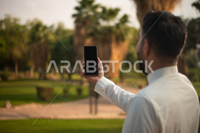 صورة من الخلف لرجل عربي خليجي سعودي يرتدي الزي السعودي التقليدي يقضي أوقات ممتعة في إحدى الحدائق في السعودية، التقاط صور تذكارية من خلال الهاتف المحمول، بث مباشر، نزهه في الهواء الطلق، جولة في الطبيعة الخضراء
