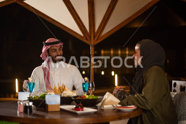 عطلة صيفية سعيدة وقضاء أوقات ممتعة ، رجل عربي خليجي سعودي يرتدي الزي السعودي التقليدي وأمرأة عربية خليجية سعودية ترتدي العباءة الخليجية السعودية يتناولان الطعام في إحدى المطاعم السعودية ،  دردشة وحوارات
