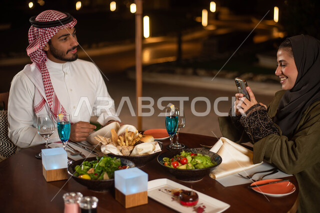 عطلة صيفية سعيدة  والتقاط صور للطعام ، رجل عربي خليجي سعودي يرتدي الثوب السعودي التقليدي والشماغ السعودي وأمرأة عربية خليجية سعودية ترتدي العباءة الخليجية السعودية يتناولان الطعام في إحدى المطاعم السعودية ، دردشة وحوارات ، قضاء أوقات ممتعة