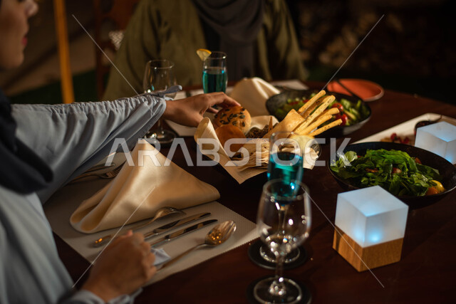 A close-up of multiple varieties of delicious food on the table inside a Saudi restaurant, two Saudi Arabian Gulf women eating delicious food in a Saudi restaurant, a plate of delicious meat with fresh bread, cold drinks and a plate of delicious green sal