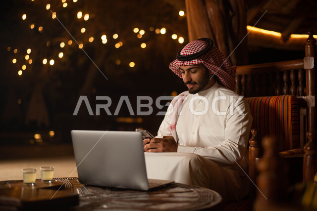  انجاز مهام العمل عبر استخدم الحاسوب المحمول ،  رجل سعودي عربي خليجي يرتدي الزي التقليدي السعودي يقضي اوقات ممتعة في احد المقاهي في السعودية ، استخدام الهاتف المحمول ، تصفح الانترنت.