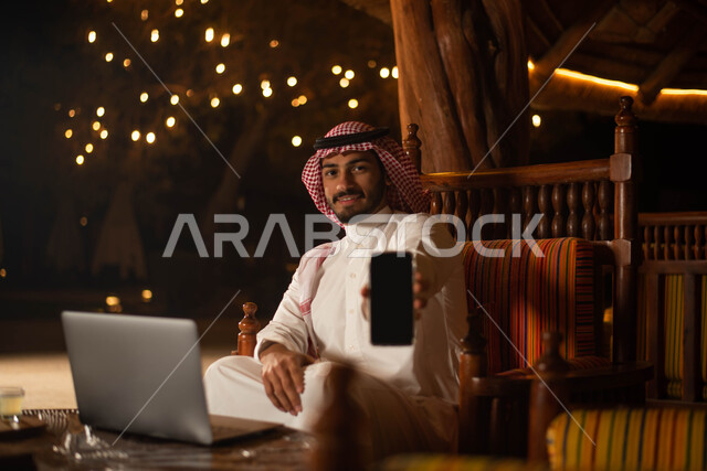 أجواء سعيدة ، تصفح الانترنت ، رجل سعودي عربي خليجي  يرتدي الزي التقليدي السعودي يقضي اوقات ممتعة في احد المقاهي في السعودية ، استخدم الهاتف المحمول بخلفية شاشة سوداء .