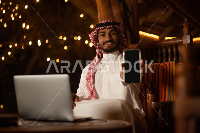  استخدم الهاتف المحمول بخلفية شاشة سوداء ،  رجل سعودي عربي خليجي يرتدي الزي التقليدي السعودي يقضي اوقات ممتعة في احد المقاهي في السعودية ، أجواء سعيدة ، تصفح الانترنت.