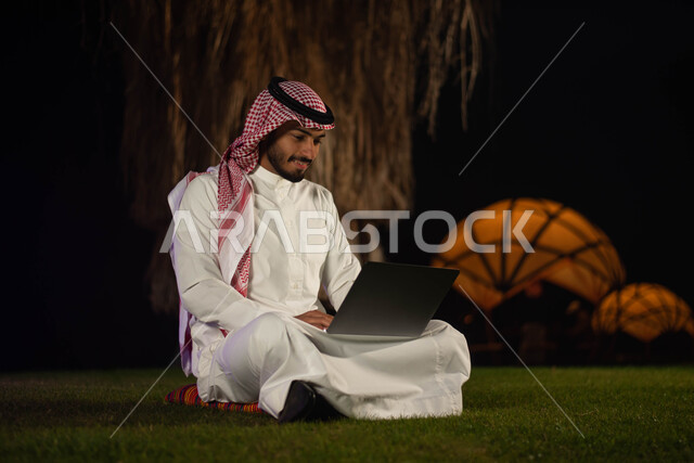  حدائق ومنتزهات ، قضاء اوقات ممتعة في الهواء الطلق ،  تصفح الإنترنت ، رجل عربي خليجي سعودي يرتدي الزي السعودي يستخدم الحاسوب المحمول ،   عقد إجتماع عمل عن بعد.