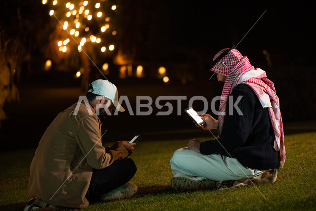 Youth session in the open air, two Saudi Arabian Gulf men using mobile phones, recreational activities, spending fun times with friends, gestures indicating joy and happiness.