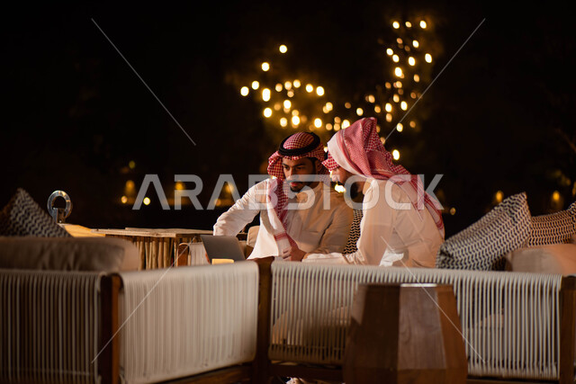 تصفح الانترنت بإستخدام أجهزة تقنية حديثة ، جلسات خارجية في الهواء الطلق ، رجلان عربيان خليجيان سعوديان يجلسان على الأريكة ، انجاز مهام العمل عبر إستخدام جهاز الحاسوب المحمول ، الحوارات والنقاشات ، اجتماع رجال الأعمال