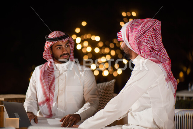 صورة مقربة لرجلان عربيان خليجيان سعوديان يجلسان على الأريكة ، انجاز مهام العمل عبر إستخدام جهاز الحاسوب المحمول ، الحوارات والنقاشات ، اجتماع رجال الأعمال ، تصفح الانترنت بإستخدام أجهزة تقنية حديثة ، جلسات خارجية في الهواء الطلق.