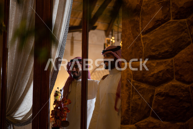 التأمل والتركيز، النظر الى الخارج من خلال النافذة ، صورة مقربة لرجلان عربيان خليجيان سعوديان يرتديان الزي السعودي التقليدي ويقفان امام النافذة، أنشطة شبابية ترفيهية ، إيماءات تدل على الفرح والسعادة