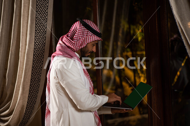 انجاز مهام ومشاريع العمل ،  رجل أعمال عربي خليجي سعودي يرتدي الزي التقليدي السعودي يحمل الحاسوب المحمول في يده  ، تصفح الإنترنت ، ادارة الأعمال عن بعد ، ايماءات تدل على السعادة والسرور.