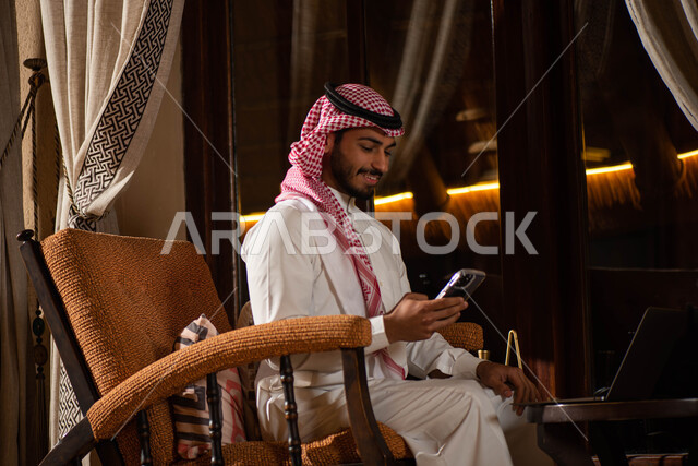  تصفح الإنترنت ، رجل أعمال عربي خليجي سعودي يرتدي الزي التقليدي السعودي يجلس على الكرسي  ، إستخدام جهاز الحاسوب المحمول لإنجاز مهام ومشاريع العمل ، ادارة الأعمال.