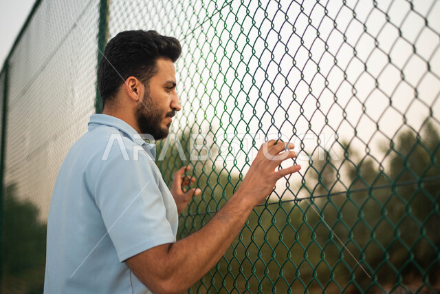 سياج شبكي، اماكن عامة، صورة من الخلف لرجل عربي خليجي سعودي يرتدي الملابس الرياضية يقف امام سياج حديد في الهواء الطلق ،  الاشجار والنباتات الخضراء