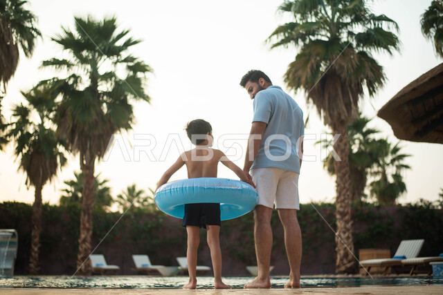 أب عربي خليجي سعودي يقضي اوقات ممتعة مع طفله، اجواء صيفية ممتعة والمشي على حدود البركة المائية ، الاستعداد للسباحة في المسبح ، رحلة سياحية ترفيهية واستخدام عوامة السباحة وقضاء عطلة صيفية سعيدة ، الاستعداد للسباحة في المسبح