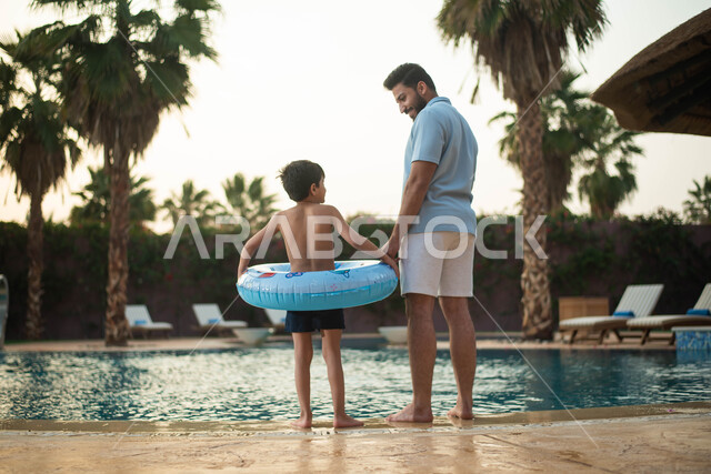 رحلة سياحية ترفيهية واستخدام عوامة السباحة وقضاءعطلة صيفية سعيدة ، أب عربي خليجي سعودي يقضي اوقات ممتعة مع طفله، اجواء صيفية ممتعة والمشي على حدود البركة المائية، الاستعداد للسباحة في المسبح