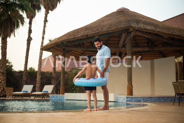 استخدام عوامة السباحة وقضاءعطلة صيفية سعيدة ، أب عربي خليجي سعودي يقضي اوقات ممتعة مع طفله، اجواء صيفية ممتعة والمشي على حدود البركة المائية، الاستعداد للسباحة في المسبح ، رحلة سياحية ترفيهية