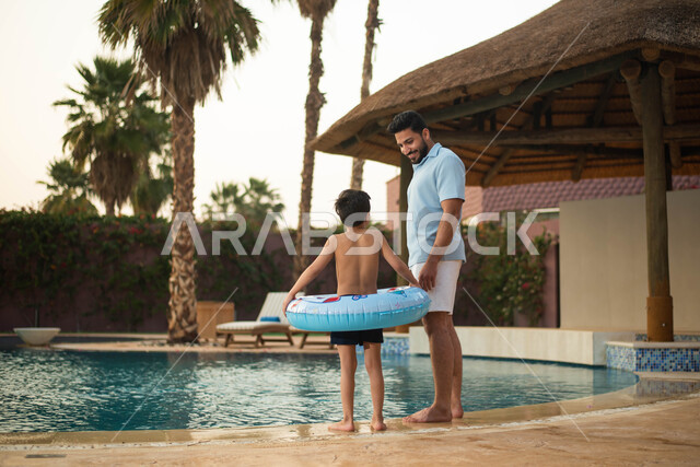 استخدام عوامة السباحة وقضاءعطلة صيفية سعيدة ، أب عربي خليجي سعودي يقضي اوقات ممتعة مع طفله، اجواء صيفية ممتعة والمشي على حدود البركة المائية، الاستعداد للسباحة في المسبح ، رحلة سياحية ترفيهية