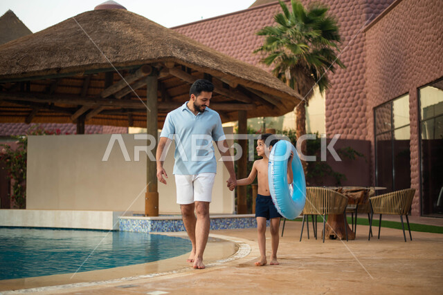 A recreational tourist trip, using a swimming buoy, spending a happy summer vacation, a Saudi Arabian Gulf man spending fun times with his son, a pleasant summer atmosphere and walking on the borders of the water pool, getting ready to swim in the pool