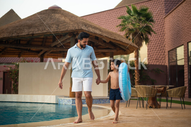 رحلة سياحية ترفيهية واستخدام عوامة السباحة وقضاءعطلة صيفية سعيدة ، رجل عربي خليجي سعودي يقضي اوقات ممتعة مع ابنه، اجواء صيفية ممتعة والمشي على حدود البركة المائية، الاستعداد للسباحة في المسبح