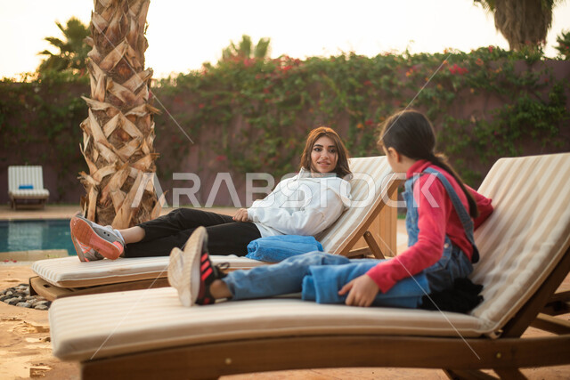 الجلوس على كرسي مريح ، شابة عربية خليجية سعودية برفقة طفلتها تجلسان على كرسي الاسترخاء على شاطيء البحر ، عطلة صيفية في منتجع سياحي ورحلة سياحية ترفيهية