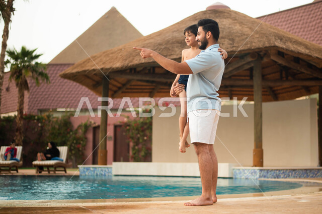 رحلة سياحية ترفيهية وعطلة صيفية سعيدة ، أب عربي خليجي سعودي يحمل ابنه ، قضاء اوقات ممتعة والاستعداد للسباحة في المسبح، اجواء صيفية ممتعة والمشي على حدود البركة المائية،  إيماءات يدين تدل على الإشارة إلى شيء ما