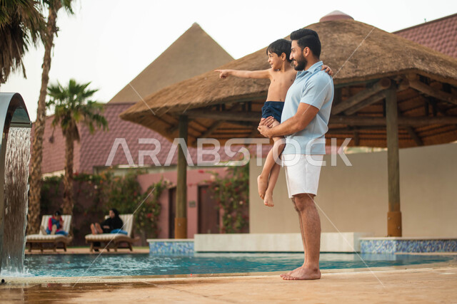 A pleasant summer atmosphere and walking on the borders of the water pool, a Saudi Arabian Gulf man carrying his son, spending fun times and getting ready to swim in the pool, an entertaining tourist trip and a happy summer vacation, two hands gestures in
