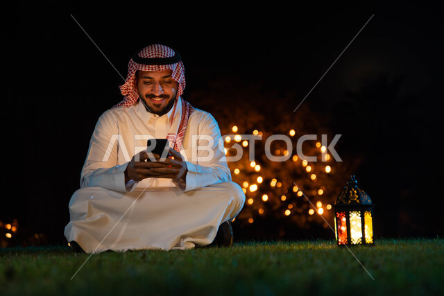 التواصل الاجتماعي وتصفح المواقع الالكترونية، شاب عربي خليجي سعودي يرتدي الزي السعودي التقليدي يستخدم الهاتف المحمول يجلس في الهواء الطلق في الخارج ، فانوس رمضان، شهر رمضان المبارك وإضاءة وزينة شهر رمضان، أجواء رمضانية