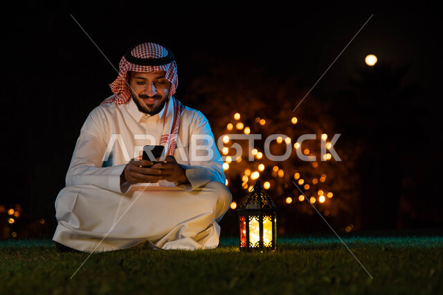 التواصل الاجتماعي وتصفح المواقع الالكترونية، شاب عربي خليجي سعودي يرتدي الزي السعودي التقليدي يستخدم الهاتف المحمول يجلس في الهواء الطلق في الخارج ، فانوس رمضان، شهر رمضان المبارك وإضاءة وزينة شهر رمضان، أجواء رمضانية