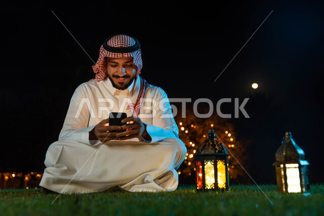 استخدام جهاز الموبايل ، رجل عربي خليجي سعودي يرتدي الزي السعودي التقليدي يستخدم الهاتف المحمول يجلس في الهواء الطلق في الخارج ، التواصل الاجتماعي وتصفح المواقع الالكترونية، فانوس رمضان، إضاءة وزينة شهر رمضان، أجواء رمضانية