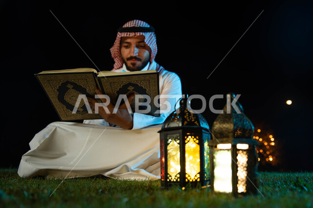 Reading, reciting and memorizing the Noble Qur’an, lighting and decorations for the month of Ramadan, a Saudi Arabian Gulf man wearing the traditional Saudi dress holding the Holy Qur’an in his hand, the blessed month of Ramadan and a Ramadan atmosphere