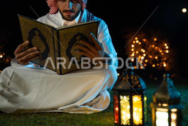Lighting and decorations for the month of Ramadan, a Saudi Arabian Gulf man wearing the traditional Saudi dress holding the Holy Qur’an in his hand, reading, reciting and memorizing the Noble Qur’an, the blessed month of Ramadan and a Ramadan atmosphere